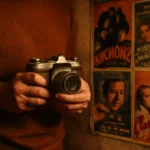 Fotografia analógica iniciantes como: fotógrafo vintage segurando câmera 35mm com parede de cartazes antigos